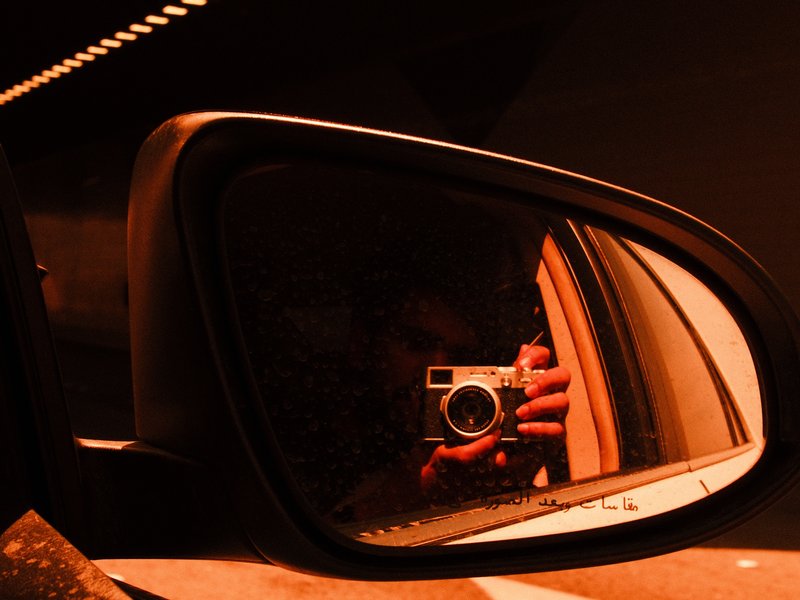 A close-up of a car side mirror reflecting a hand holding a camera in warm orange tones.