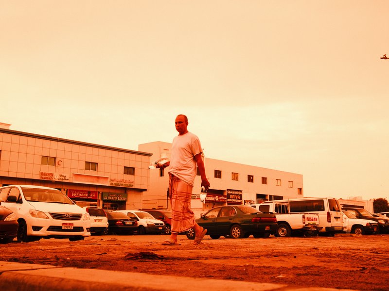 A man walking on a sandy street with vehicles parked in the background under an orange sky.