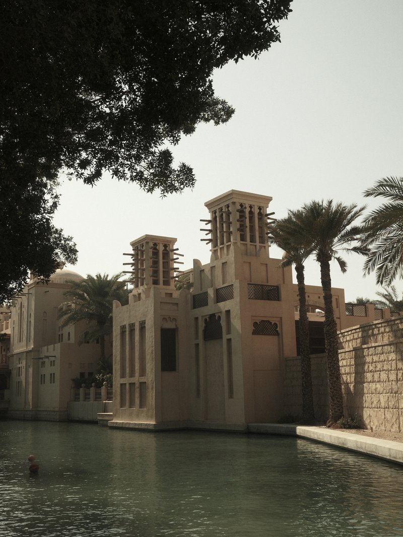 A traditional Middle Eastern building with wind towers beside a calm waterway, surrounded by palm trees.