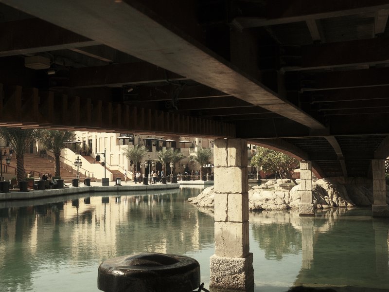 View from under a bridge showing a calm waterway with reflections and palm trees in the background.