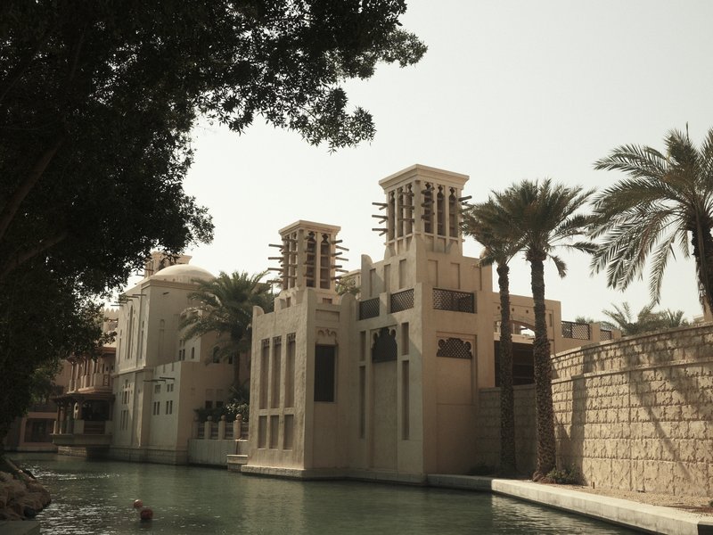 A view of a traditional Middle Eastern building beside a calm waterway, surrounded by palm trees.