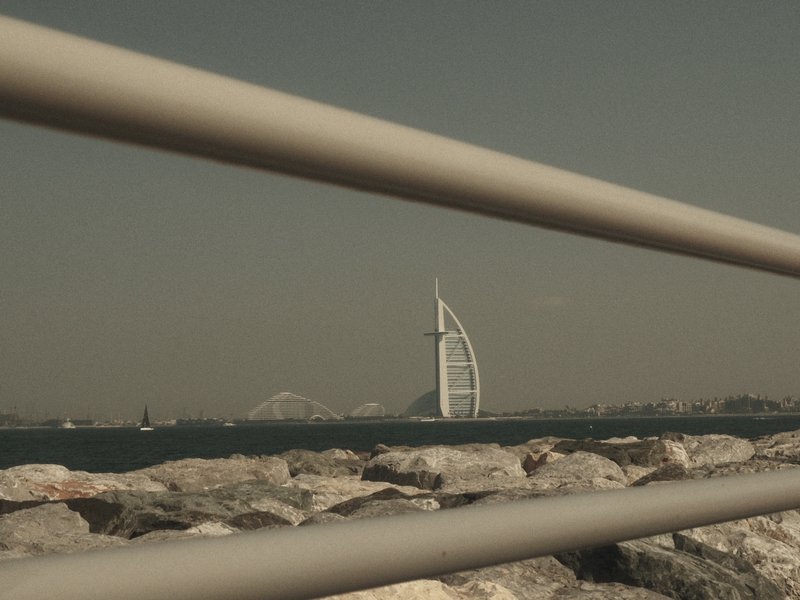 View of the Burj Al Arab hotel framed by a railing, with rocky shoreline and distant mountains.