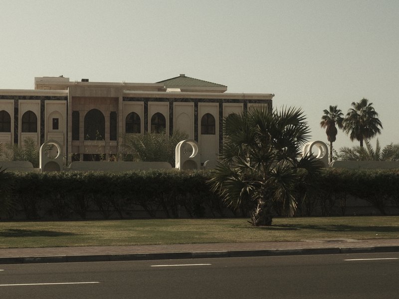 A large, elegant building with palm trees and sculptures in the foreground, set against a clear sky.