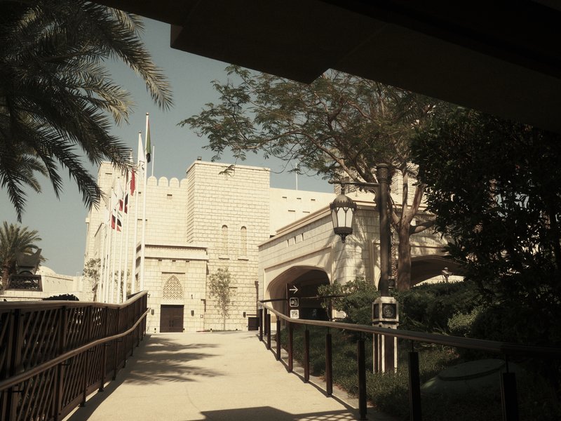 A historic building with palm trees and flags, viewed from a walkway under a shaded area.