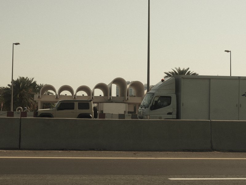 A truck and a yellow vehicle on a highway with a building featuring arches in the background.