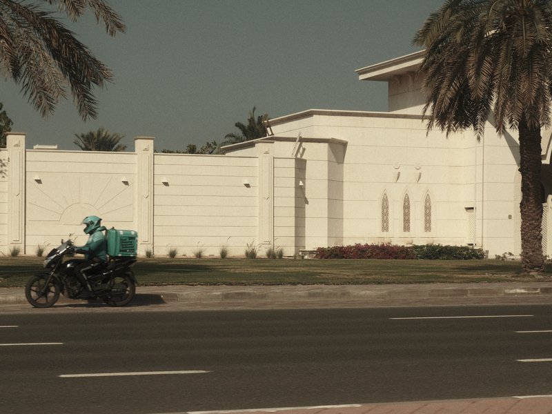 A delivery rider on a motorcycle passes by a modern white building surrounded by palm trees.