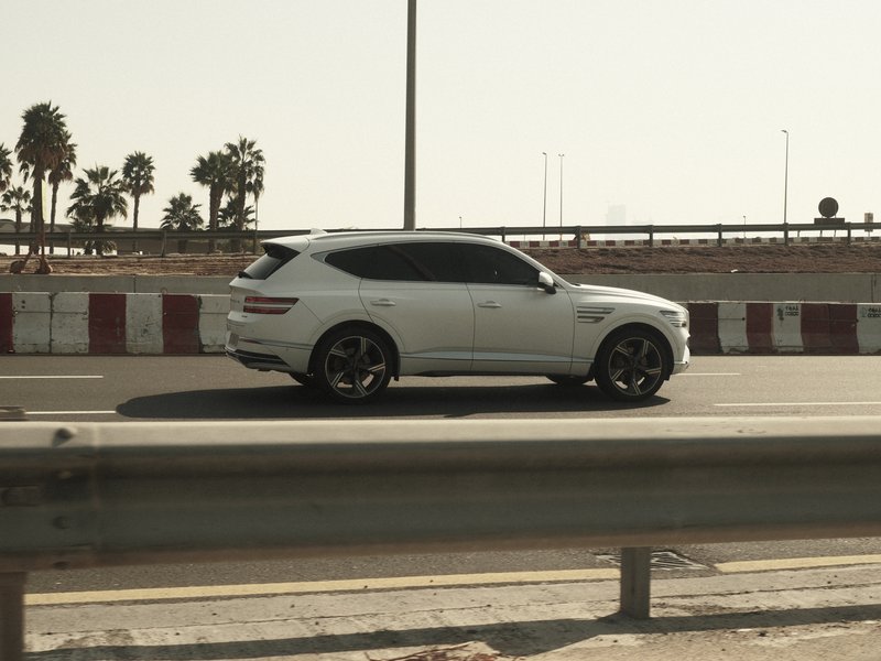 A white SUV driving on a highway with palm trees in the background under bright sunlight.