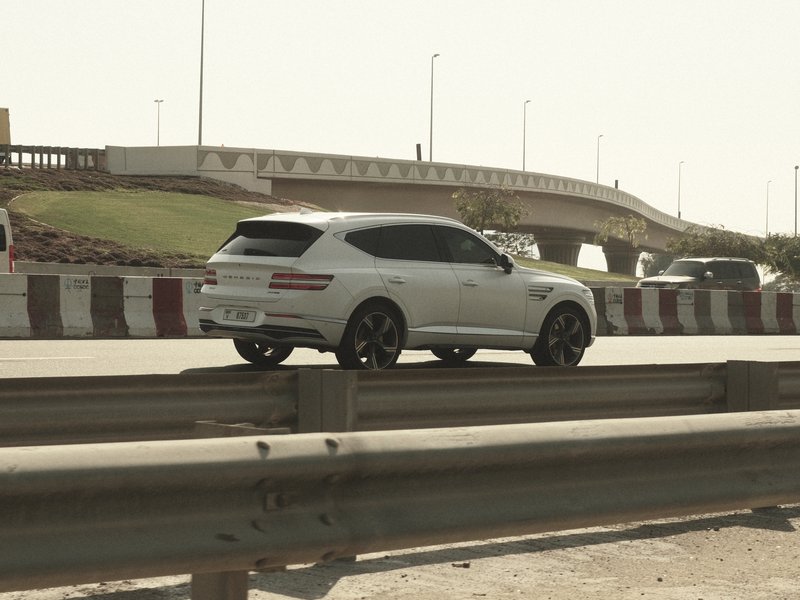 A white SUV driving on a highway with a bridge in the background under bright sunlight.