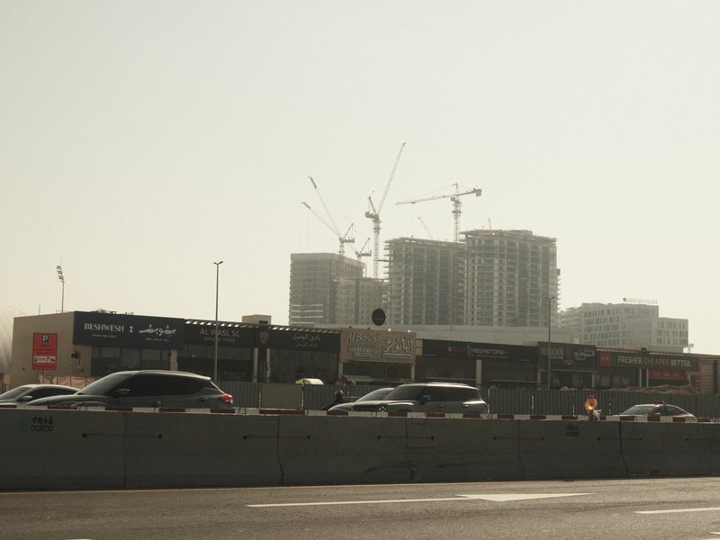 A busy urban scene with cars on a road and construction cranes in the background under a hazy sky.