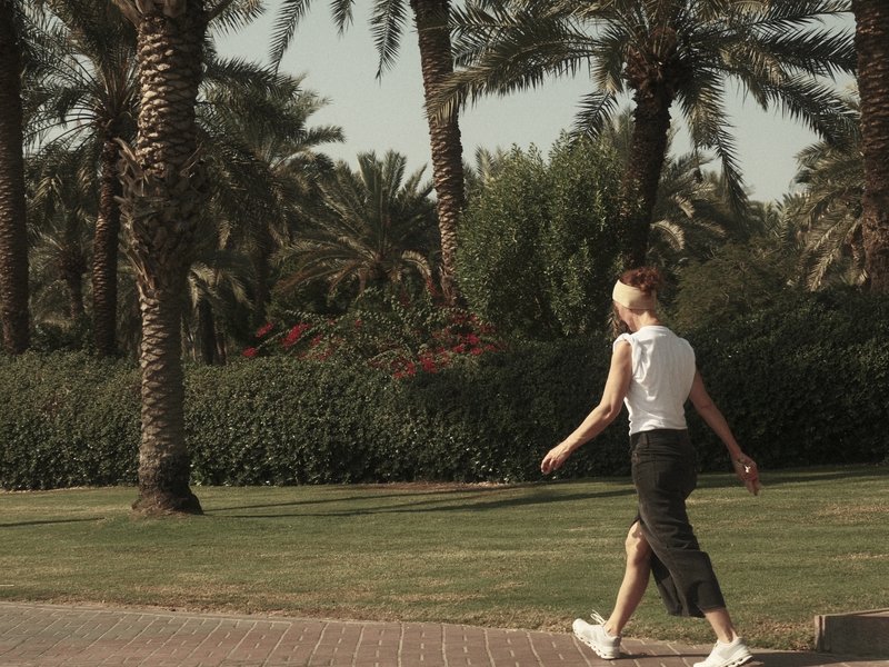 A woman walking on a path lined with palm trees and greenery in a sunny outdoor setting.