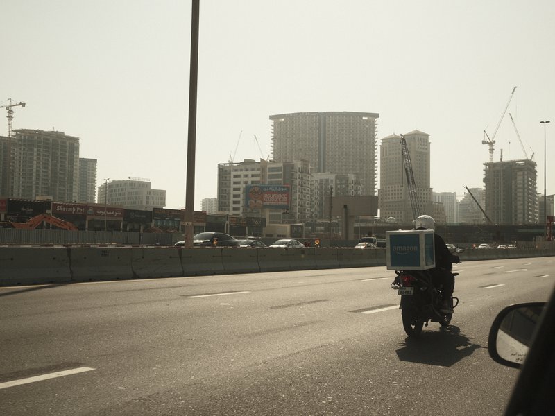 A motorcycle courier with a large delivery box rides on a busy highway with tall buildings in the background.