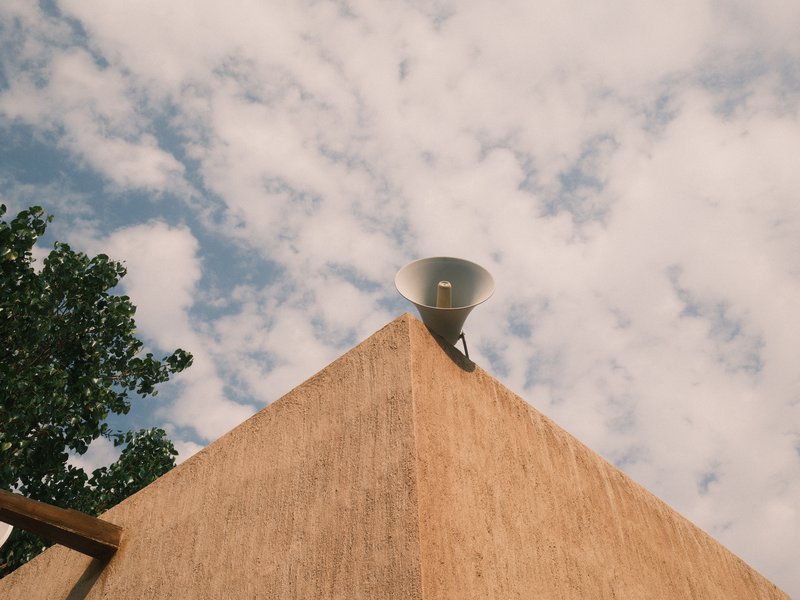 A close-up view of a building's corner with a speaker on top against a cloudy sky.