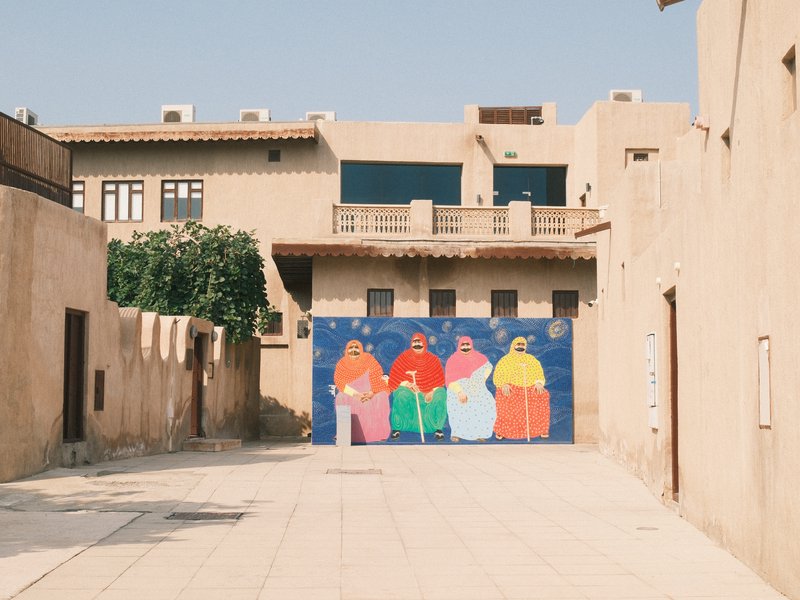 Colorful mural of four figures on a wall in a narrow street with traditional buildings.