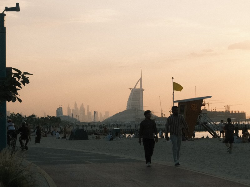 Two people walking along a beach promenade at sunset with a city skyline in the background.