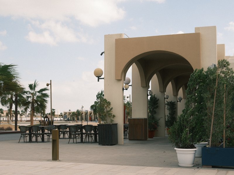 A coastal promenade featuring arches and potted plants, with tables and chairs in the background.