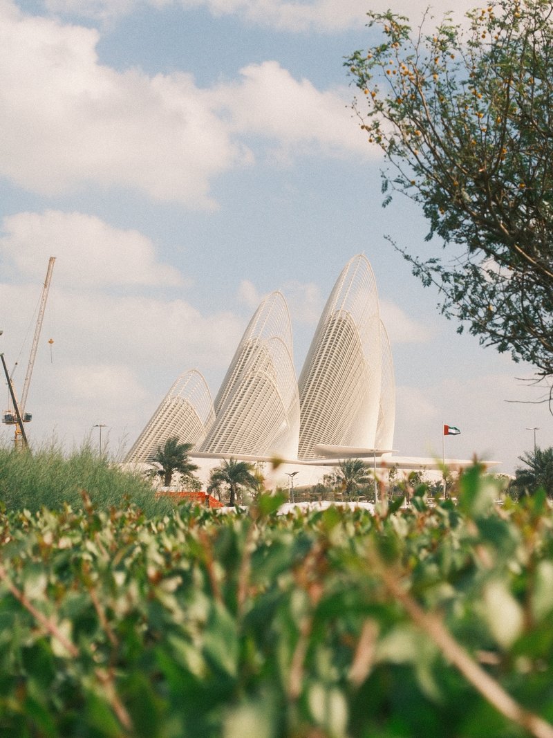 A modern architectural structure with curved white forms rises above green foliage under a blue sky with clouds.