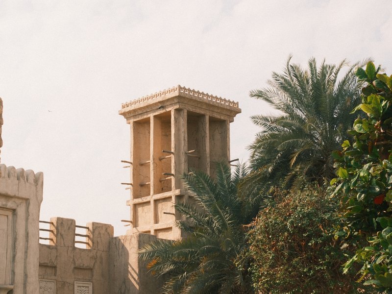 A traditional Middle Eastern building with a wind tower surrounded by palm trees under a bright sky.