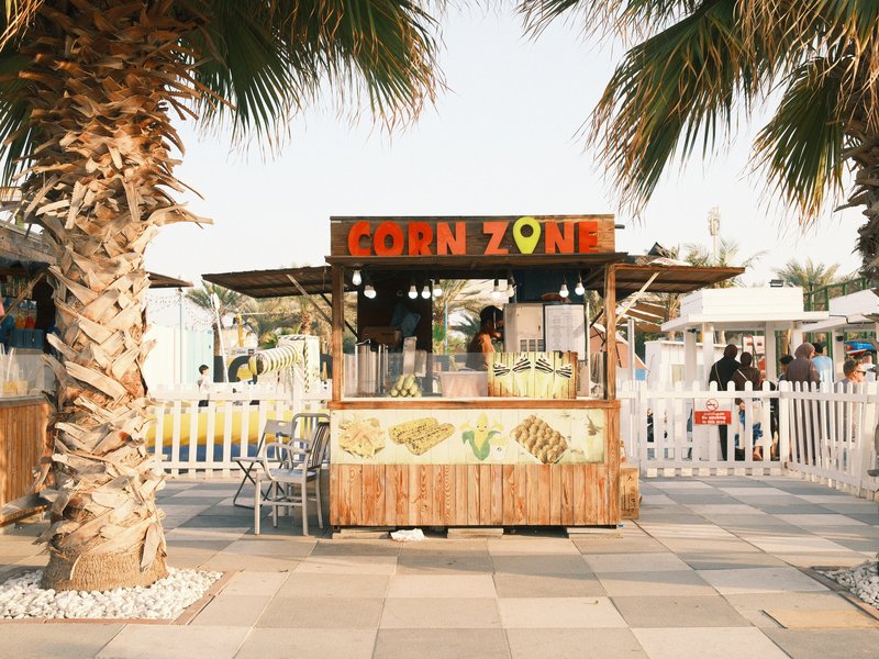 A vibrant corn stand with a sign reading 'CORN ZONE' surrounded by palm trees and a lively atmosphere.