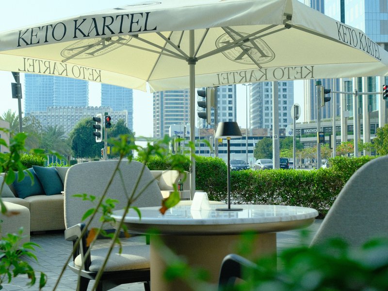 Outdoor cafe setting with tables, umbrellas, and city buildings in the background.