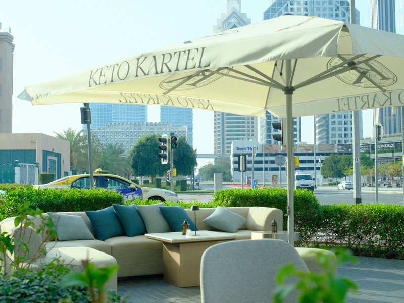 Outdoor seating area with a large umbrella, surrounded by greenery and city buildings in the background.