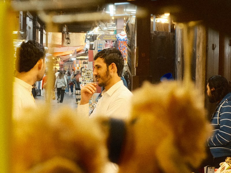 Two men engaged in conversation in a bustling market, with soft focus on teddy bears in the foreground.