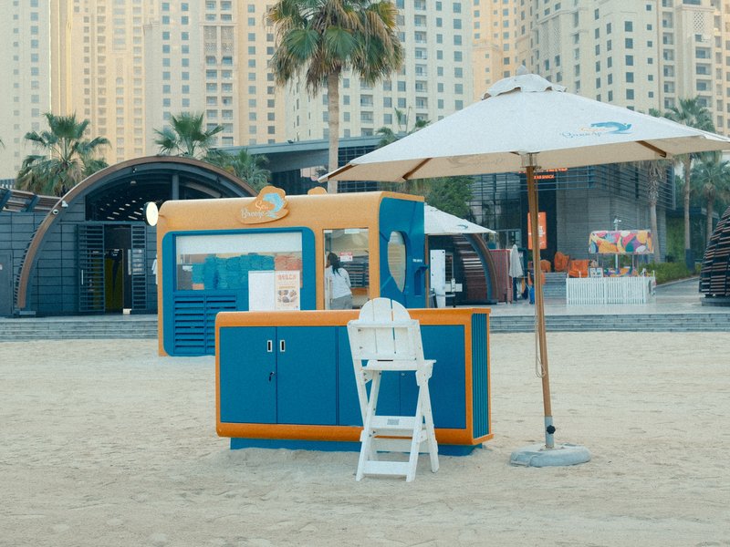 A colorful beach kiosk with an umbrella on sandy ground, surrounded by palm trees and tall buildings.