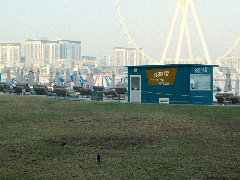 A beachside kiosk with a blue exterior near lounge chairs and a large Ferris wheel in the background.