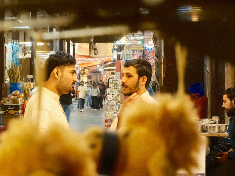 Two men engaged in conversation in a bustling market, with colorful stalls in the background.