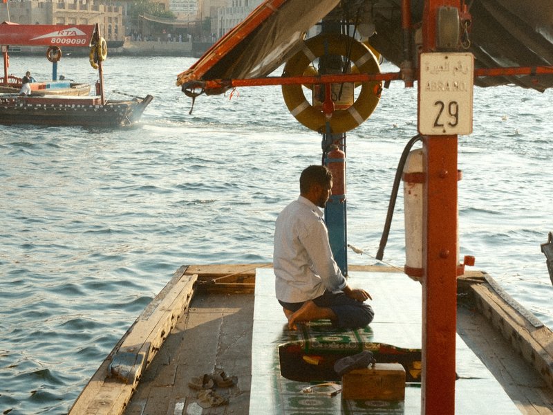 A man sitting on a boat docked by the water, with another boat in the background and a cityscape visible.