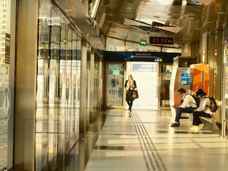 A modern train station corridor with glass walls, a woman walking, and two people sitting.