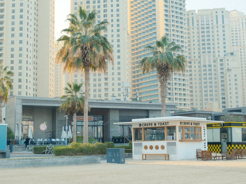 A beach scene with palm trees, a food stall, and tall buildings in the background.