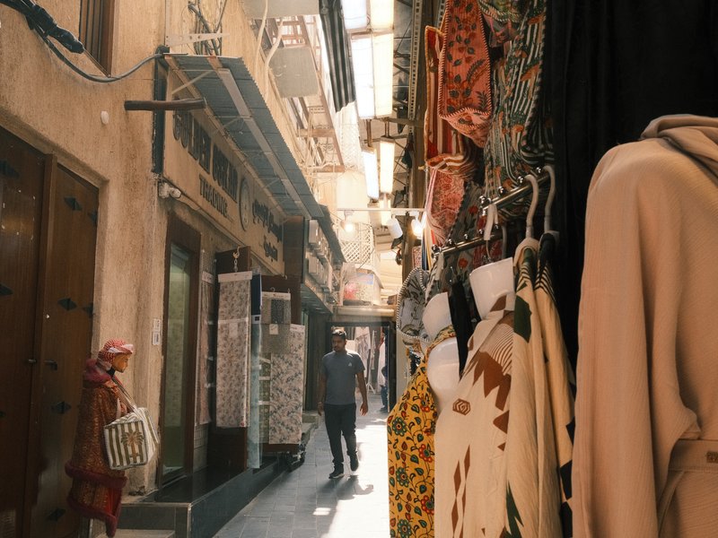 Narrow alleyway with colorful clothing hanging, a person walking, and a woman carrying a basket.