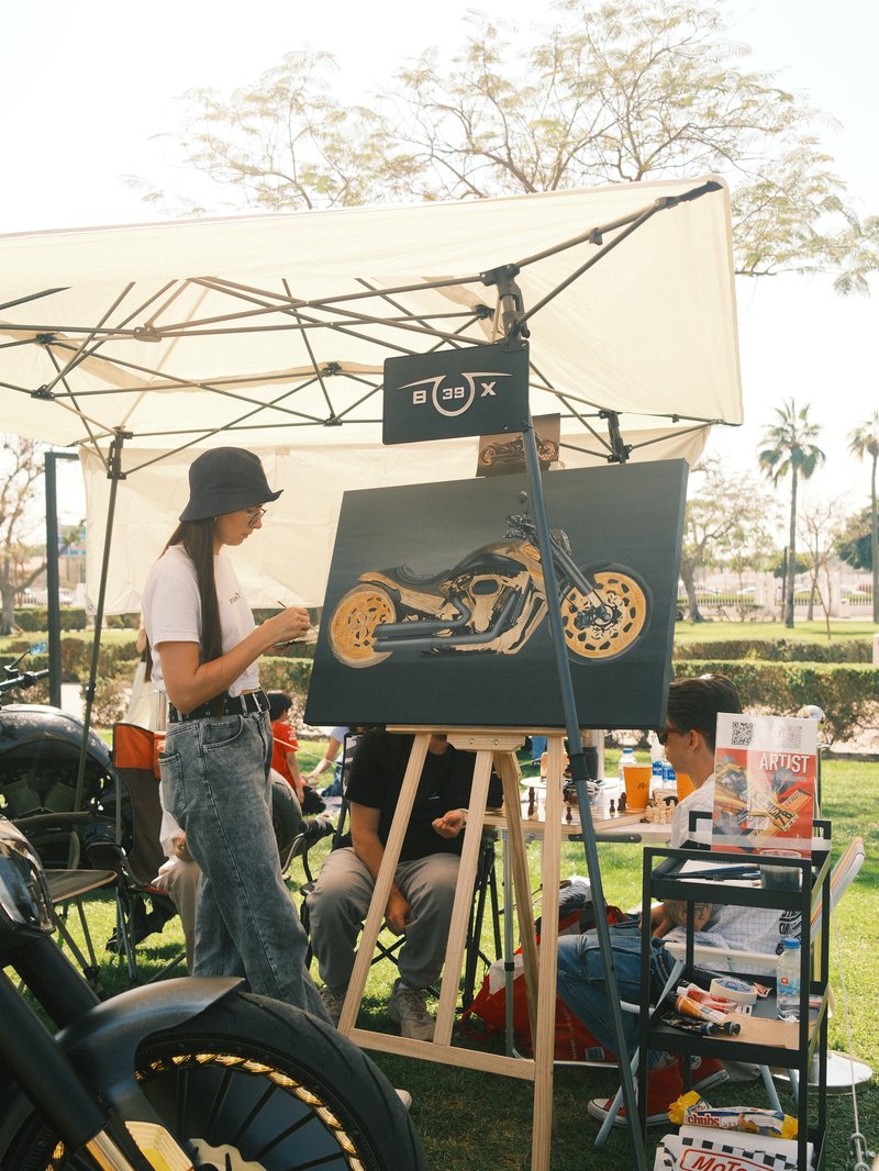 A woman painting a motorcycle artwork at an outdoor event under a tent.