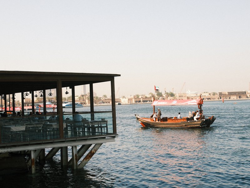 A wooden boat on water near a waterfront restaurant with outdoor seating.