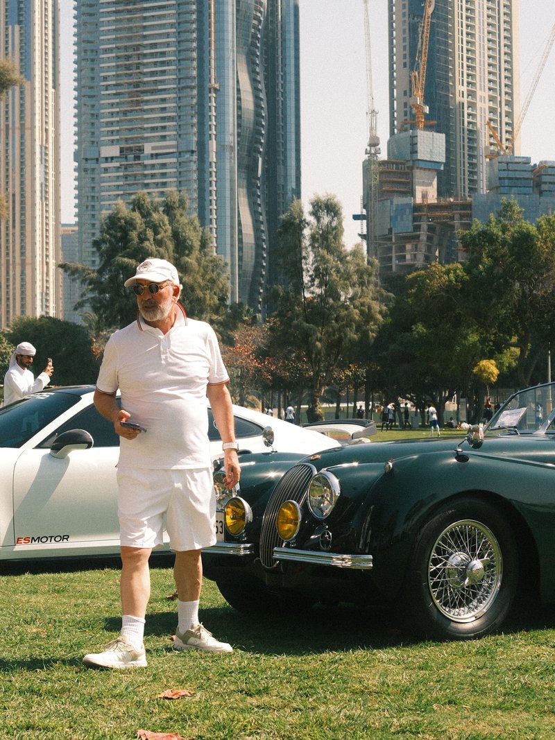A man in white attire stands beside a classic car in an urban park with skyscrapers in the background.