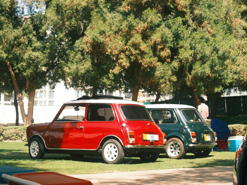 Two vintage cars, a red one and a green one, parked on grass under trees with a building in the background.