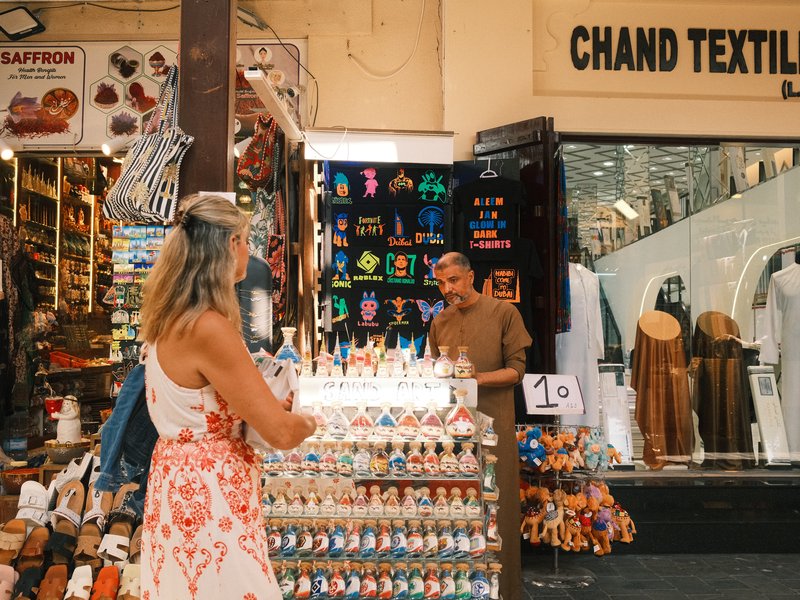 A woman in a white dress shops at a vibrant market stall filled with colorful items.