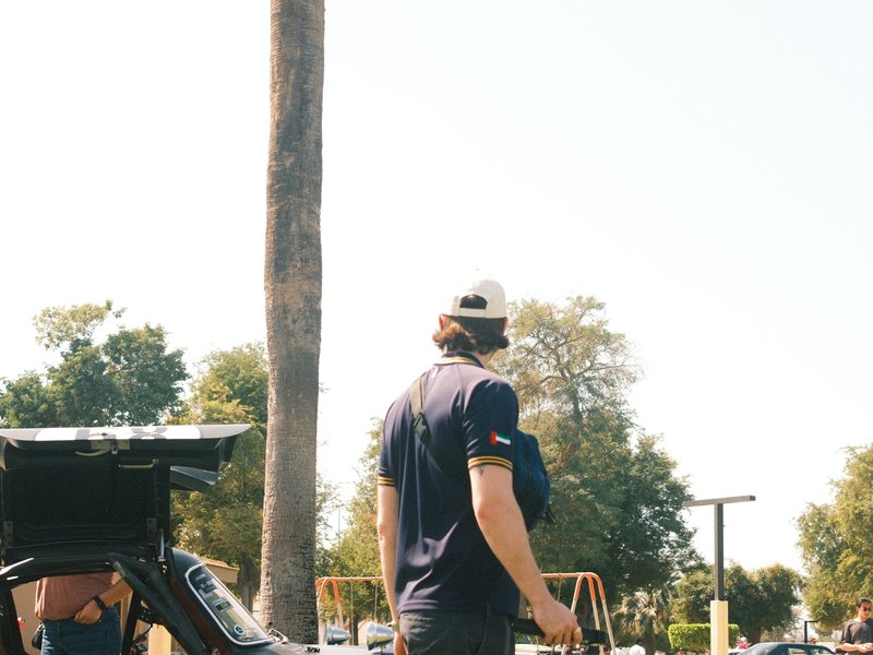 A person with a cap stands near a palm tree in a park, with swings visible in the background.