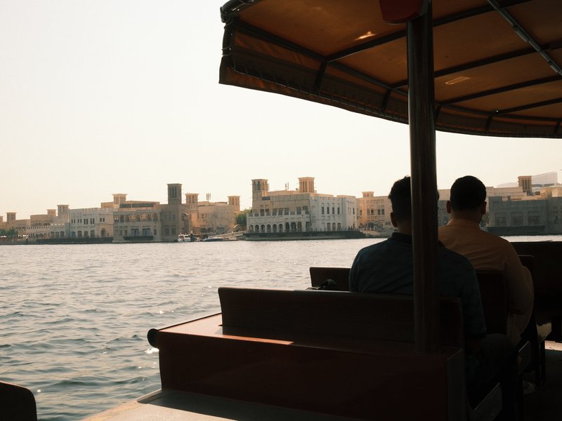 Two people sitting on a boat with a view of a coastal city in the background under bright sunlight.