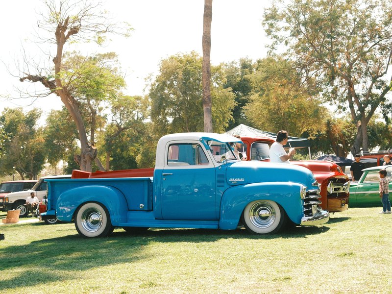 A vintage blue pickup truck parked on green grass at a car show with trees in the background.