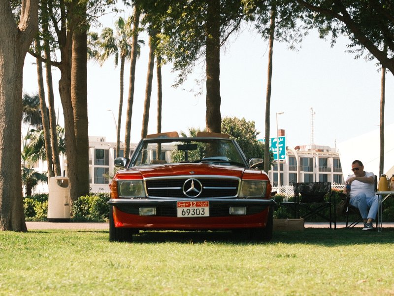 A vintage red Mercedes convertible parked on green grass with palm trees in the background.