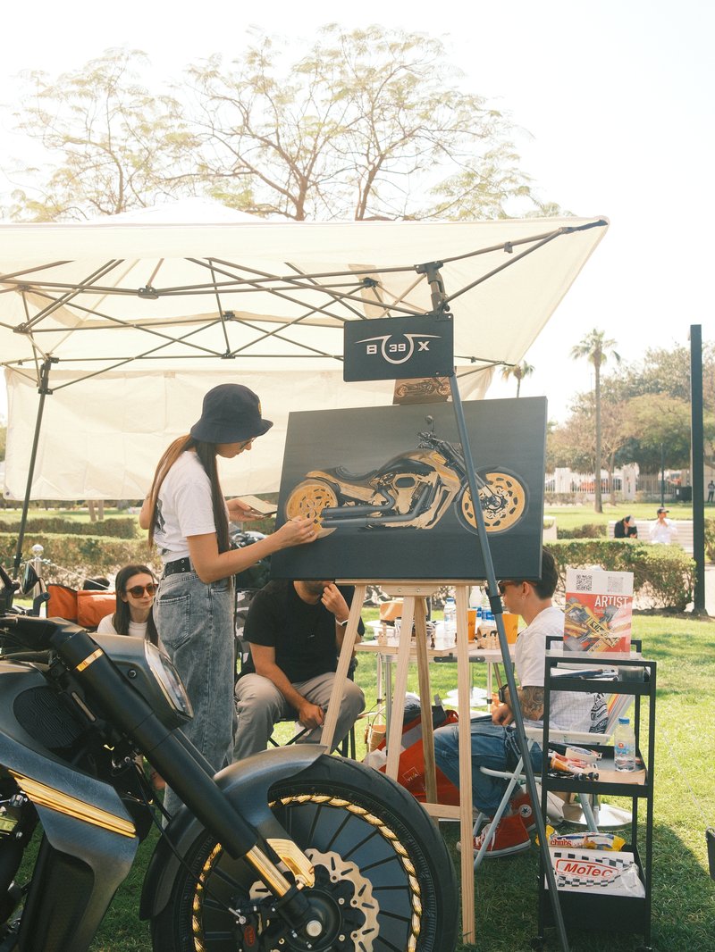 A woman painting a motorcycle on a canvas under a tent at an outdoor event.
