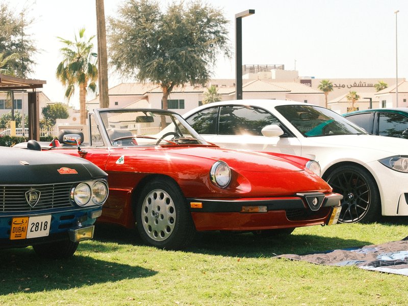 A red convertible sports car parked on green grass with two other cars nearby under bright sunlight.