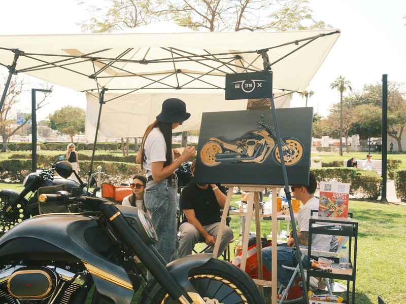 A woman painting a motorcycle art piece at an outdoor event under a canopy.