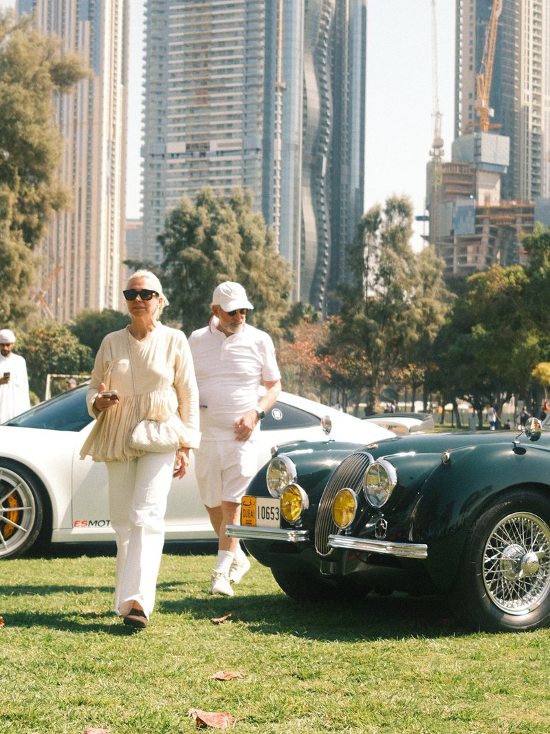 Two people walking in a park with modern skyscrapers in the background and classic cars nearby.