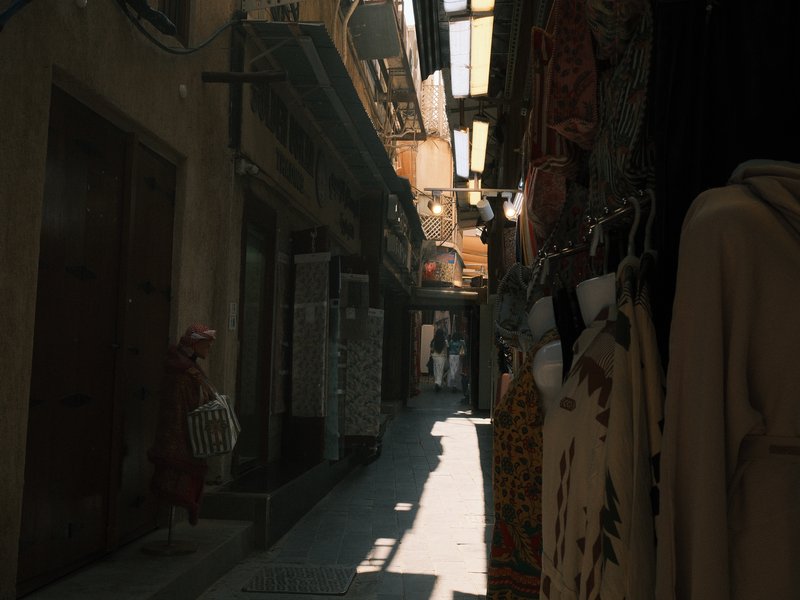 Narrow alleyway with shops on either side, sunlight streaming through overhead structures.
