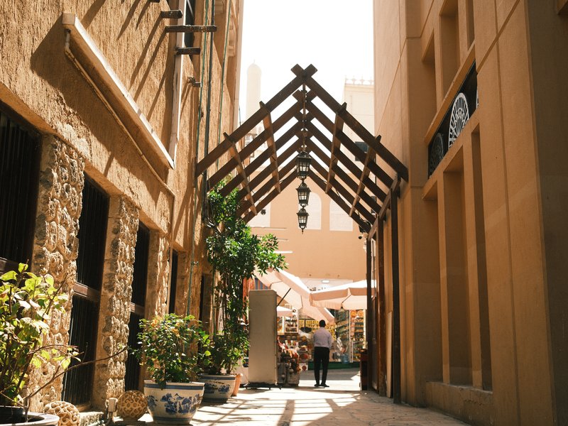 A sunlit alleyway with a wooden archway, leading to a cafe with outdoor seating.