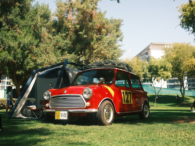 A vintage red car parked on green grass with trees in the background and a tent nearby.
