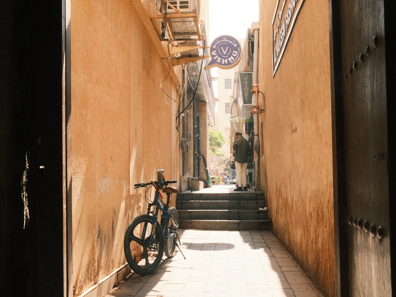 Narrow alleyway with a bicycle leaning against a wall, warm yellow tones and a person in the distance.