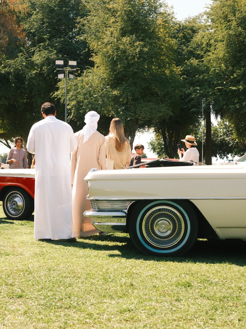 Group of people in traditional attire admiring classic cars at an outdoor event on a sunny day.